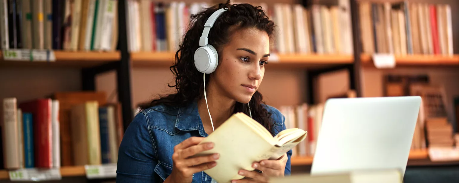 An English major holds a book and studies in a library while wearing a headset on her ears.