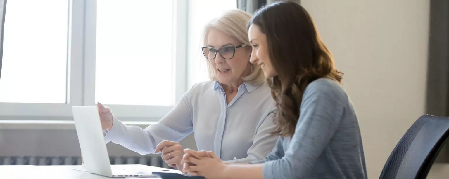 A manager points on something on a laptop screen as she delivers positive employee feedback to a smiling, receptive member of the team.