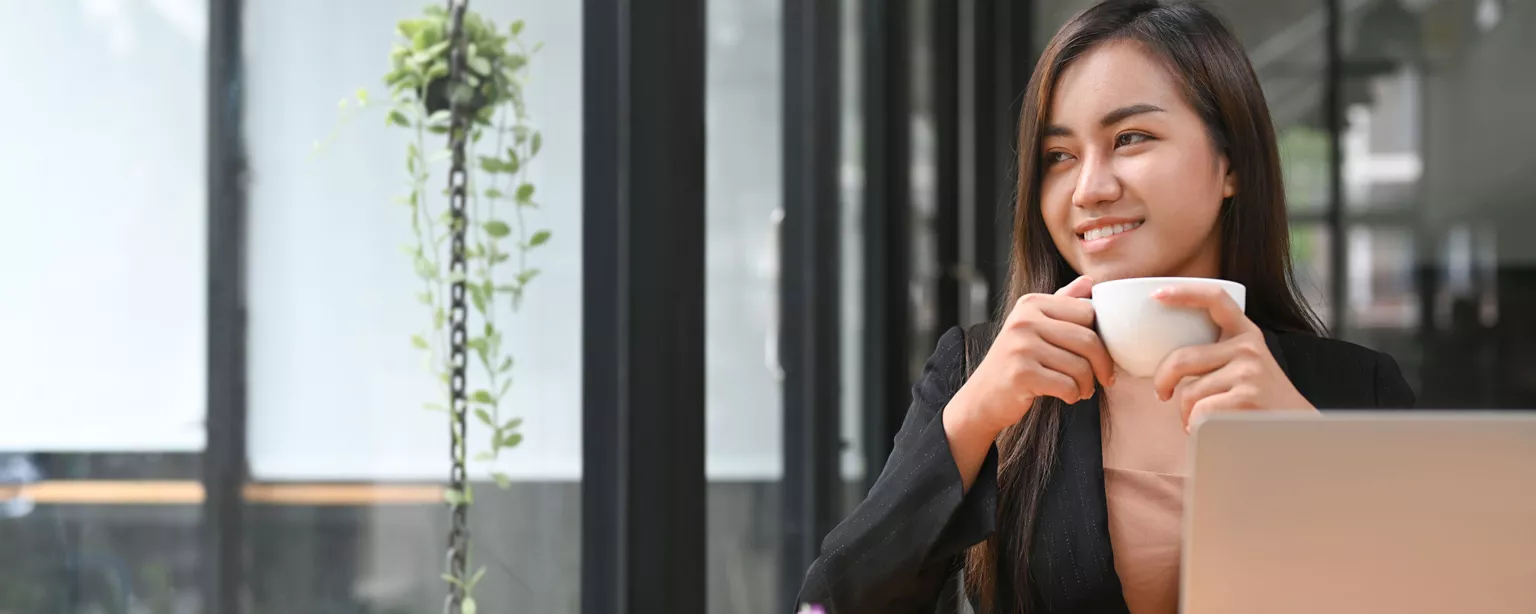 A young woman in business attire smiles while holding a coffee cup at her desk in a modern, well-lit office.