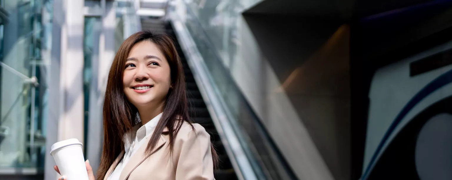 A smiling woman at the bottom of an escalator holds a coffee cup and laptop.