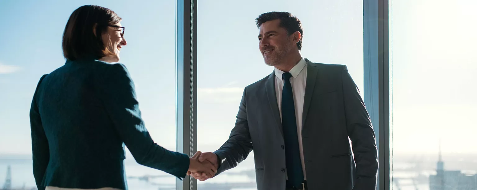 A newly hired executive shakes hands with another member of the team in a high-rise office with a view of a city in the background.