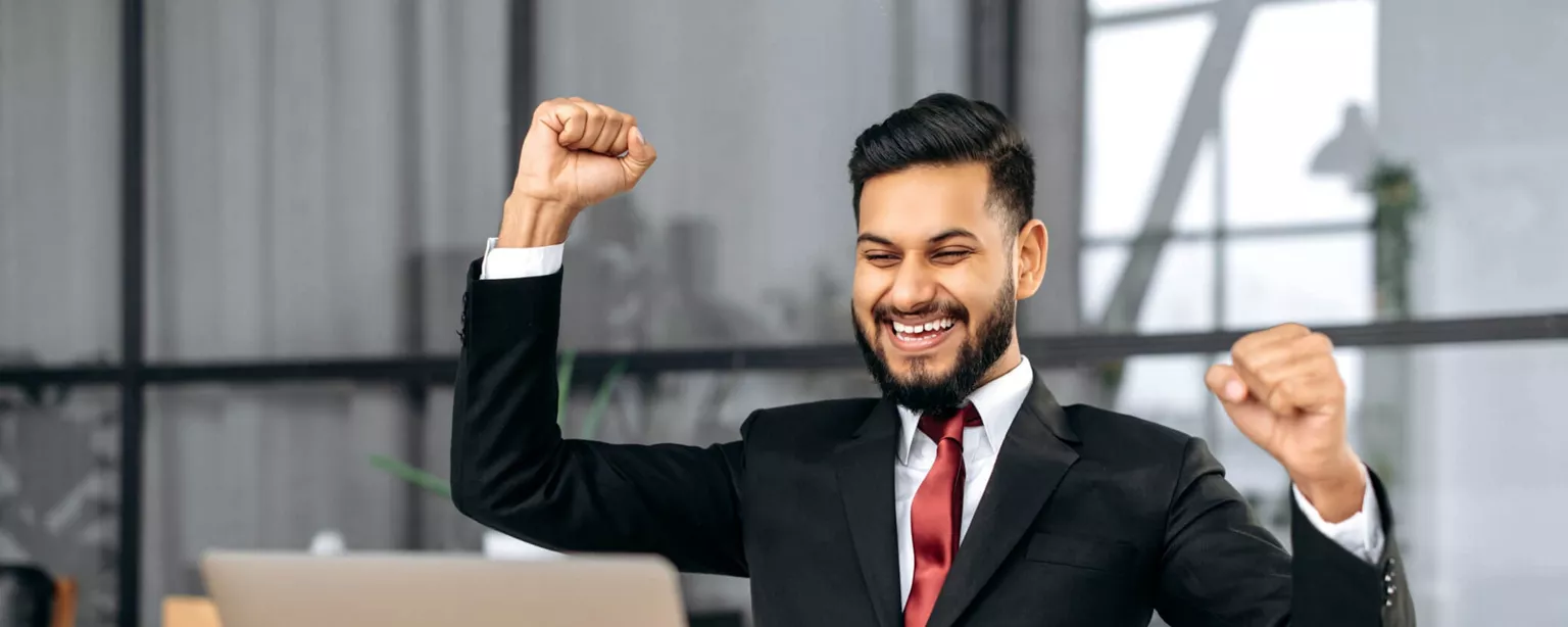 A young executive with a beard, wearing a business suit, sits with arms raised in front of a laptop and smiles because he just received good news.