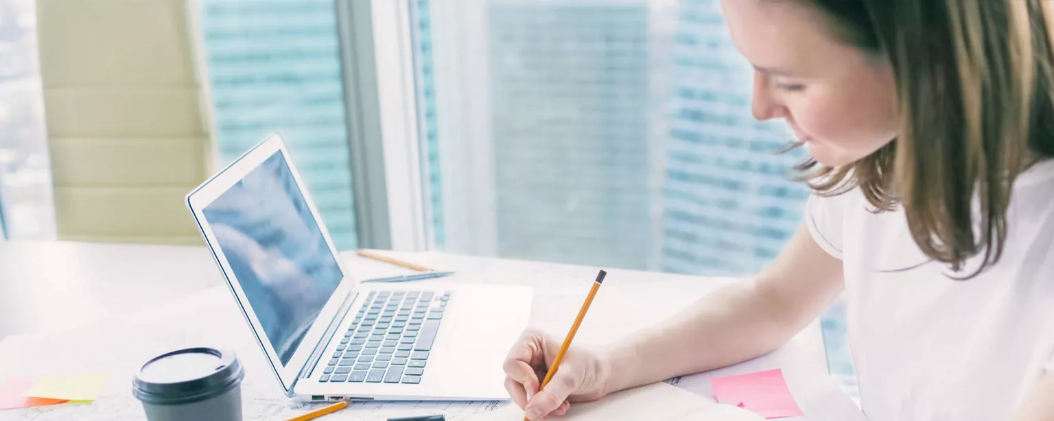 A woman works at a desk with pencil, papers, coffee cup and laptop.