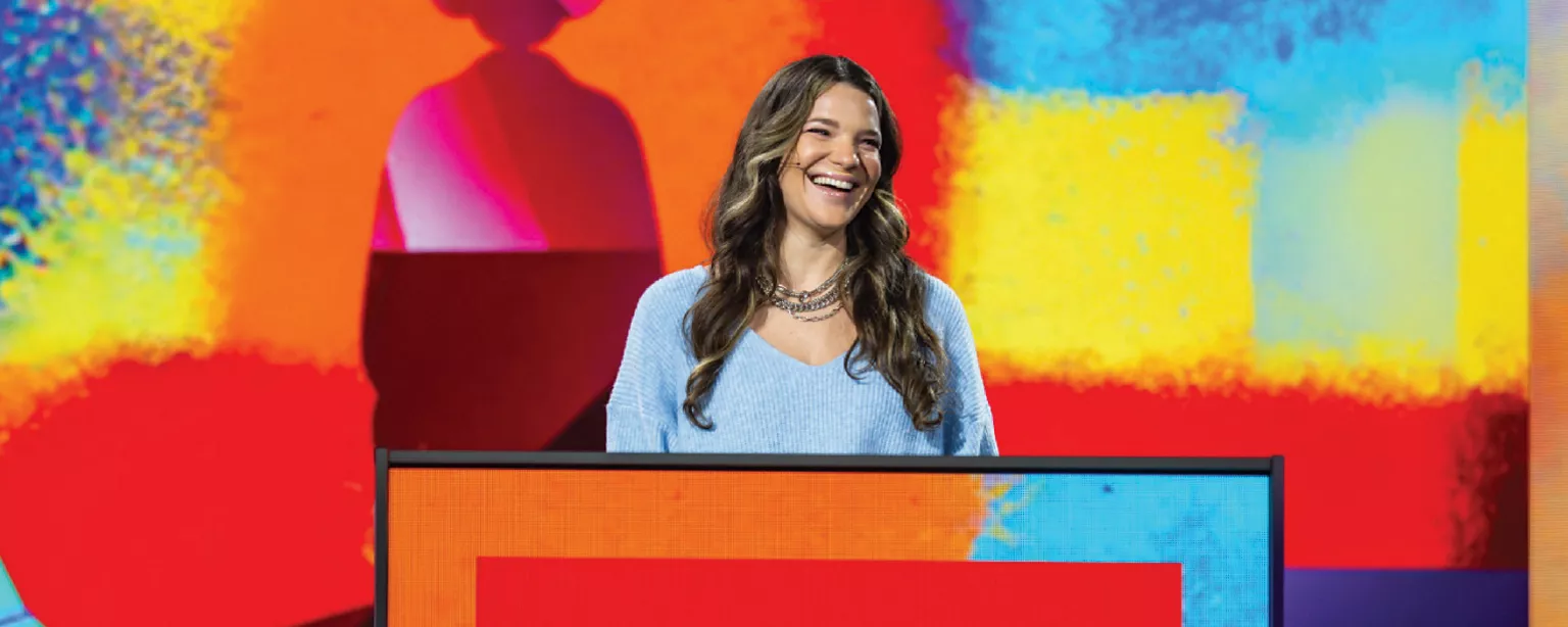 A smiling woman stands behind a podium on a brightly colored stage for Adobe Max 2025.