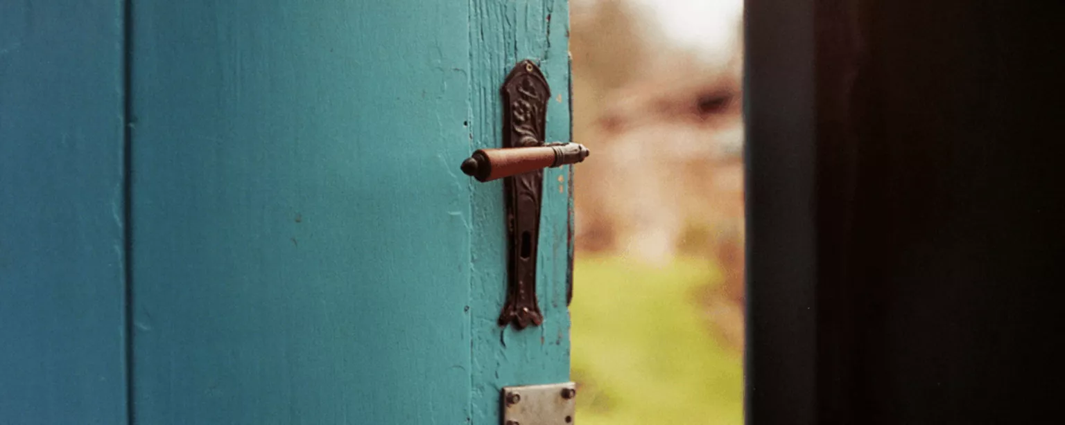 Blick ins Freie durch den Spalt einer alten, halb geöffneten Holztür.