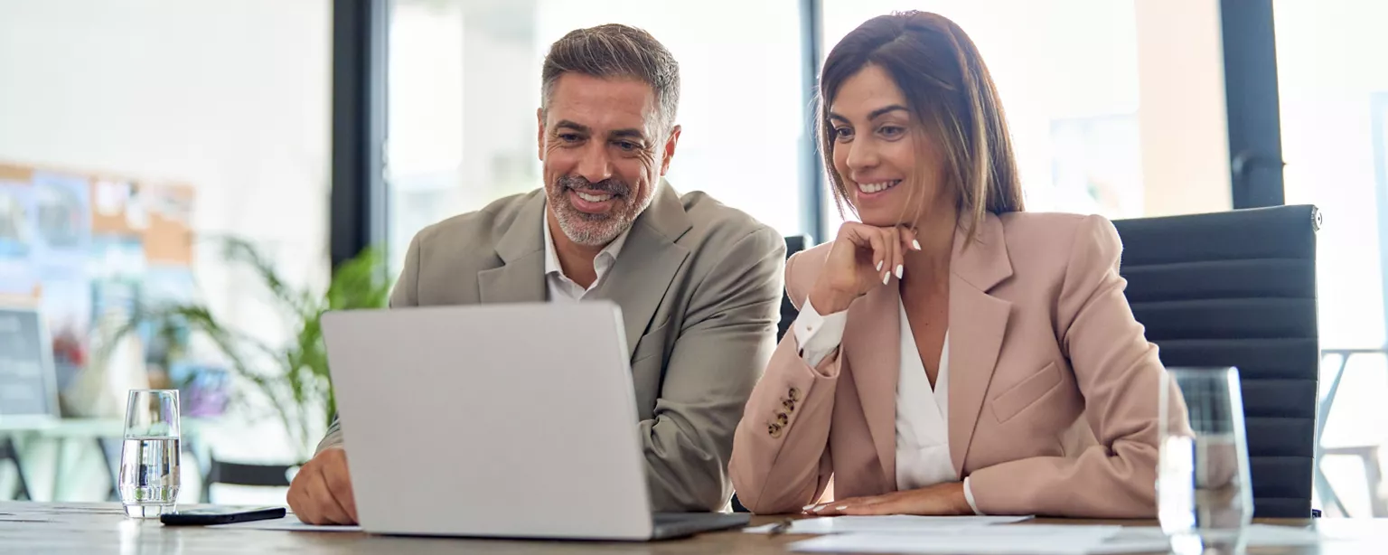 A middle-aged male and female coworker sit at a desk smiling while looking at a laptop.