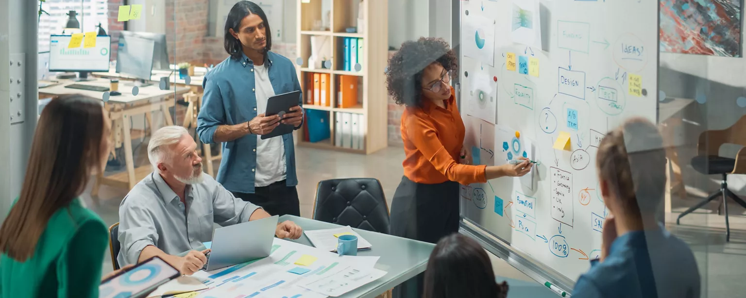 A diverse group of five creatives brainstorm near a whiteboard as a woman writes down their ideas.