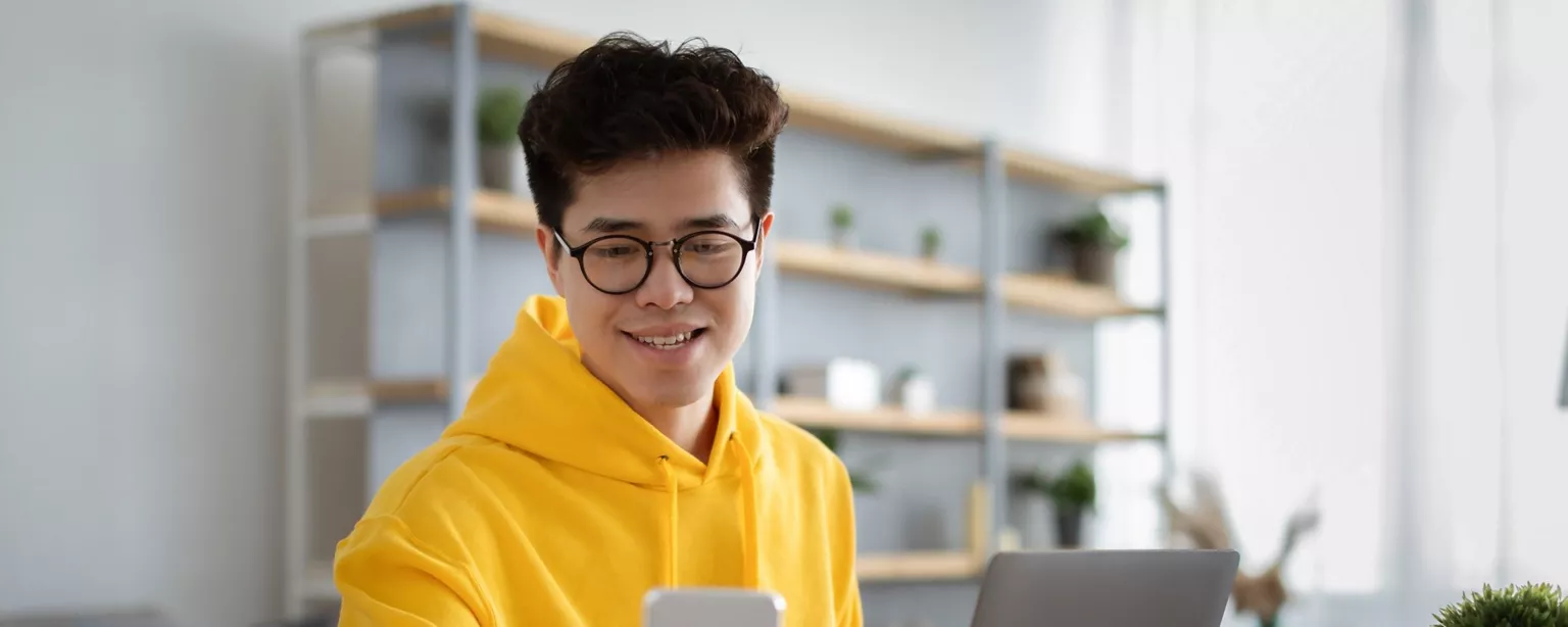 Student wearing yellow sweatshirt with phone and laptop at desk
