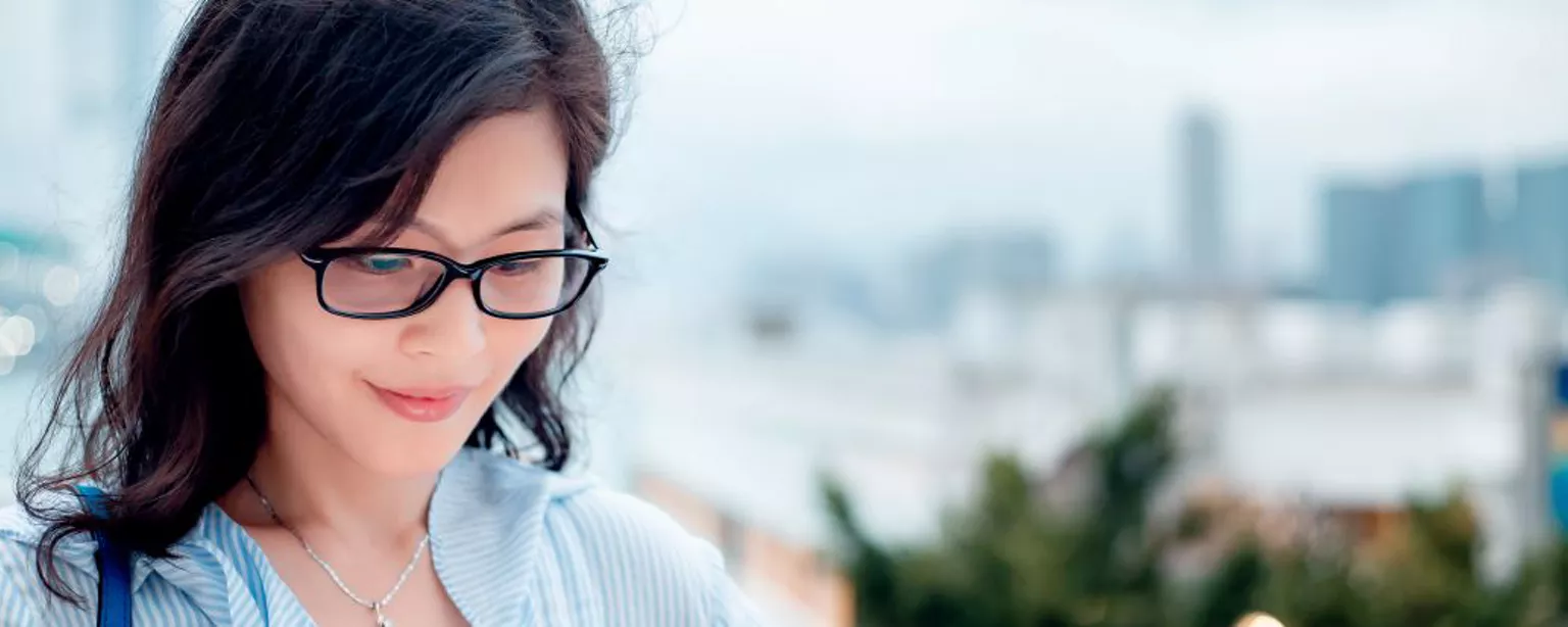 Young woman with glasses standing outside and looking at her smartphone and smiling.