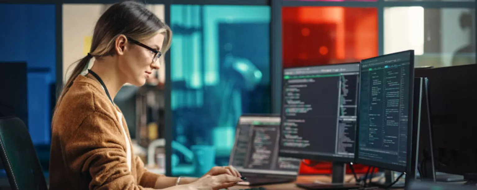 A young woman with glasses and a ponytail who works in the tech field is sitting at a desk in a modern office and looking at code on a computer screen.