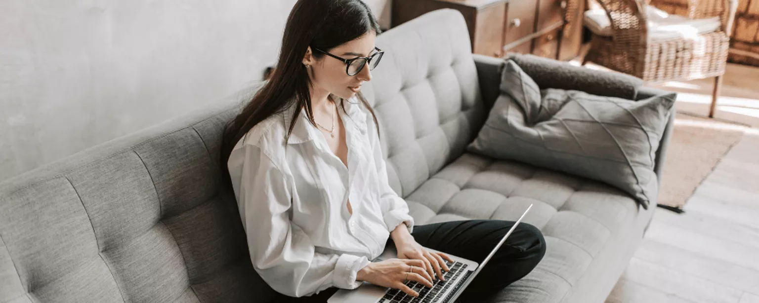 Eine junge Frau sitzt lässig auf dem Sofa und arbeitet auf ihrem Notebook, welches sie auf dem Schoß hat.