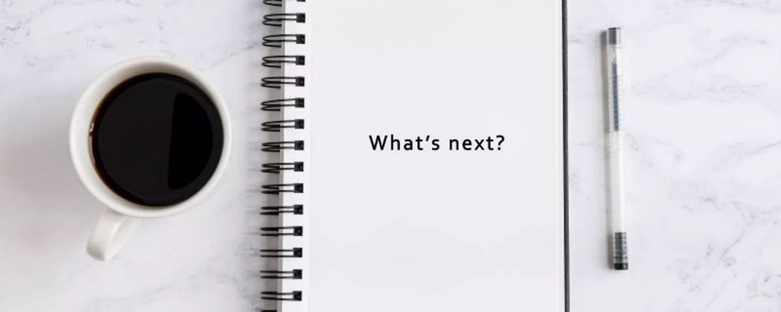 A cup of black coffee and a pen are placed next to notepad that reads, "What's next?" 
