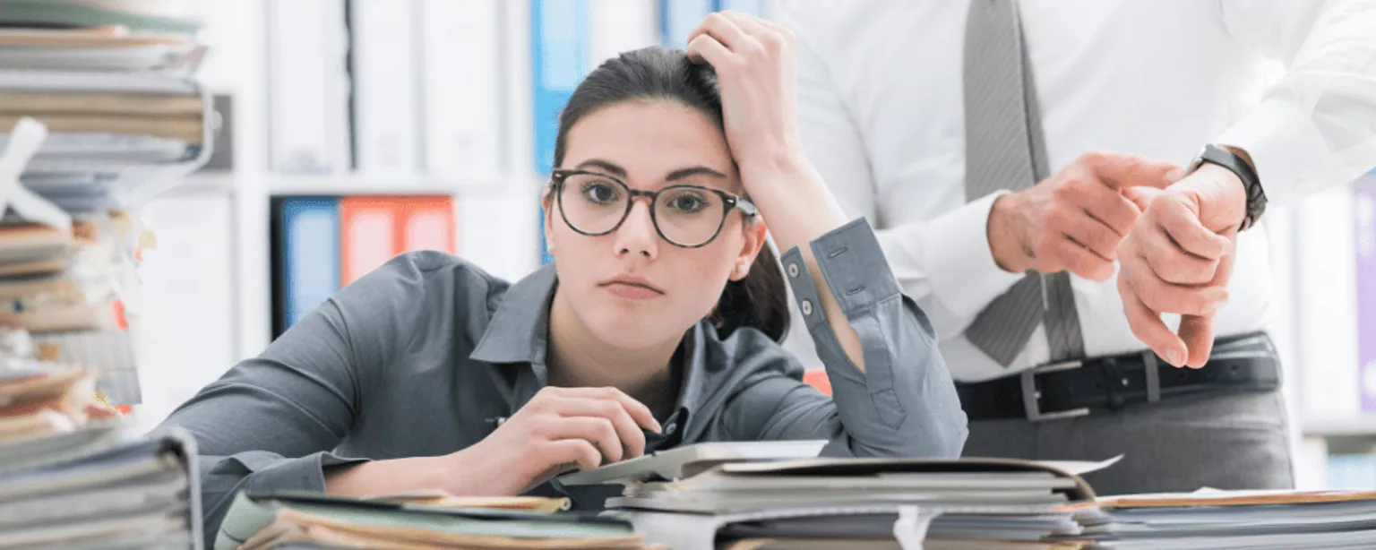 Eine Frau lehnt sich erschöpft an vielen Unterlagen ab. Hinter ihr steht ein Mann und zeigt auf die Uhr.