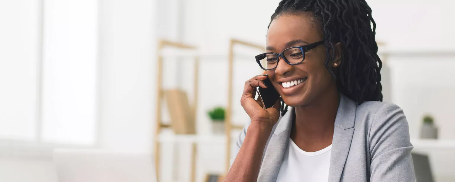 Young woman with glasses sitting at a desk and smiling while looking at a laptop and talking on a cell phone.