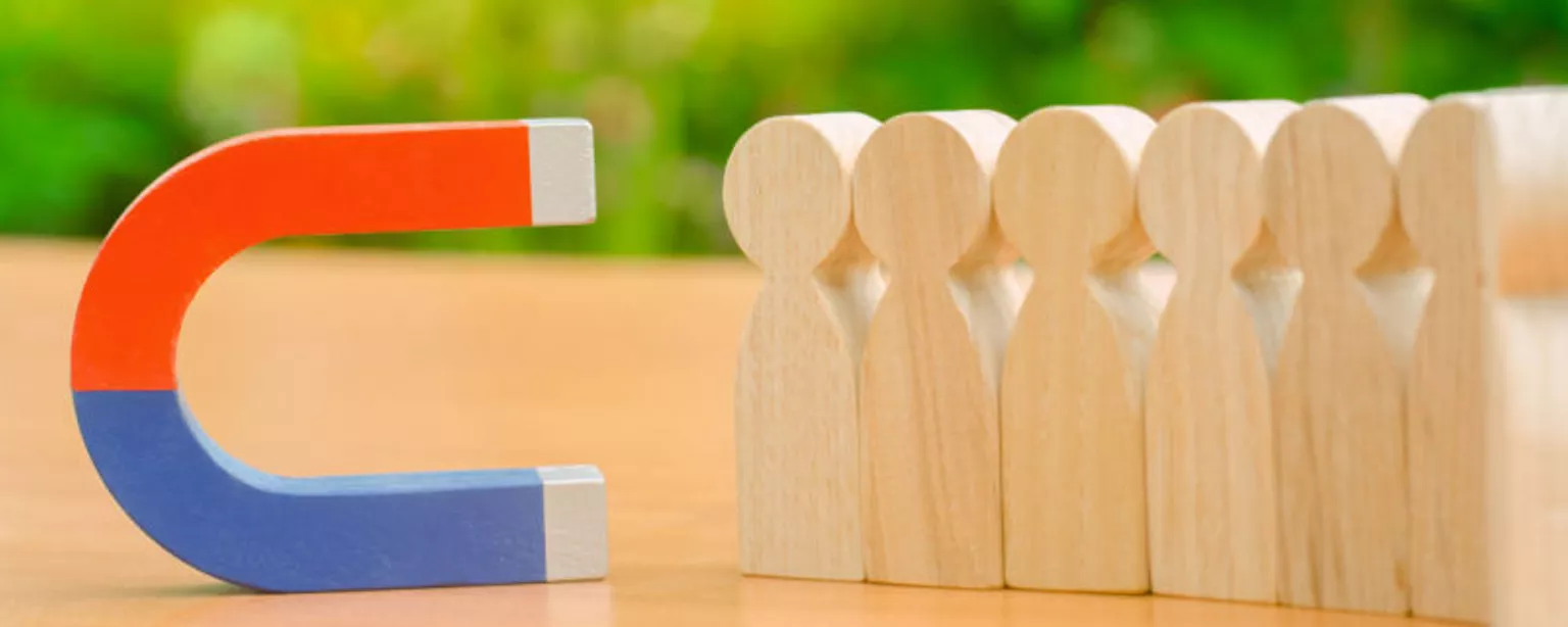 A red and blue magnet is positioned next to a row of wooden blocks that look like people, conveying a concept of "attracting talent."