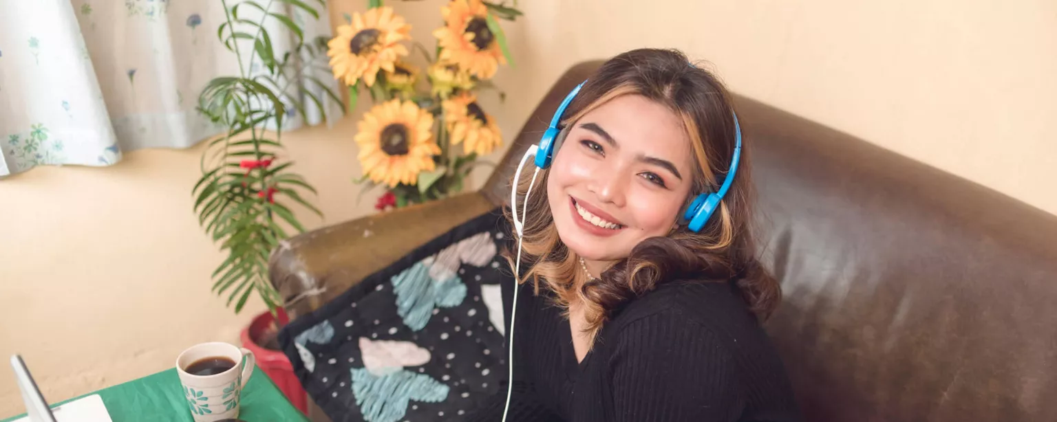 Customer services rep working at home, wearing blue headphones, smiling and sitting on a couch with a laptop in front of her.