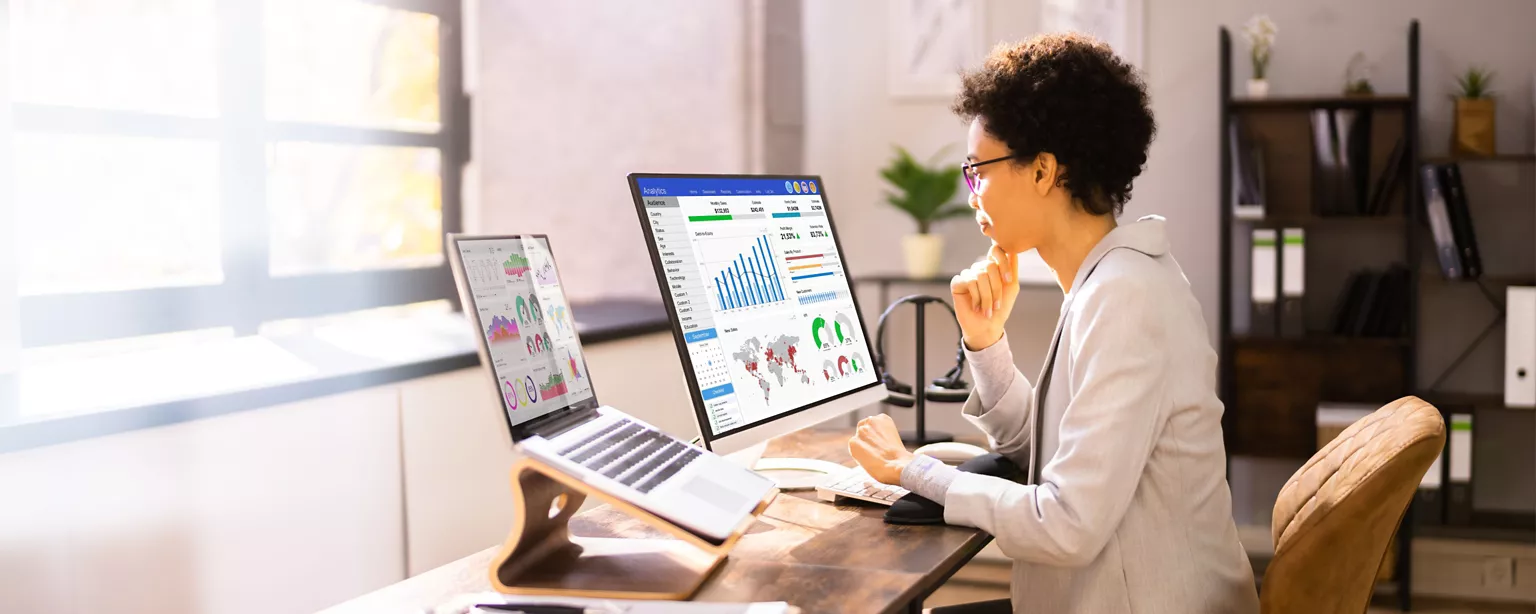 A financial analyst is sitting at a desk and looking at two computer screens displaying financial charts and other data.