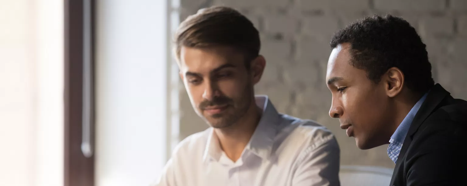 A male boss and male employee are sitting side by side at a table with a laptop in front of them; the boss is gesturing to the laptop screen and talking..