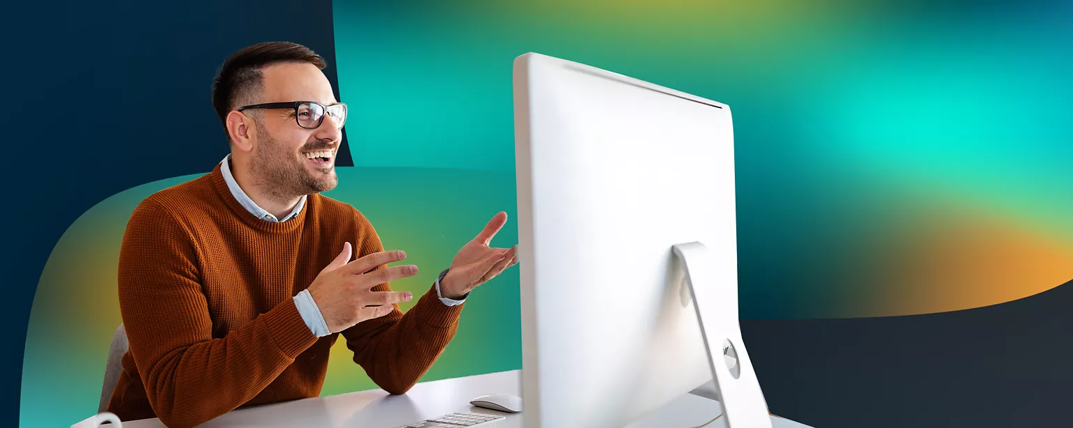A man wearing glasses and a brown sweater sits at a desk in front of a large computer monitor smiling.