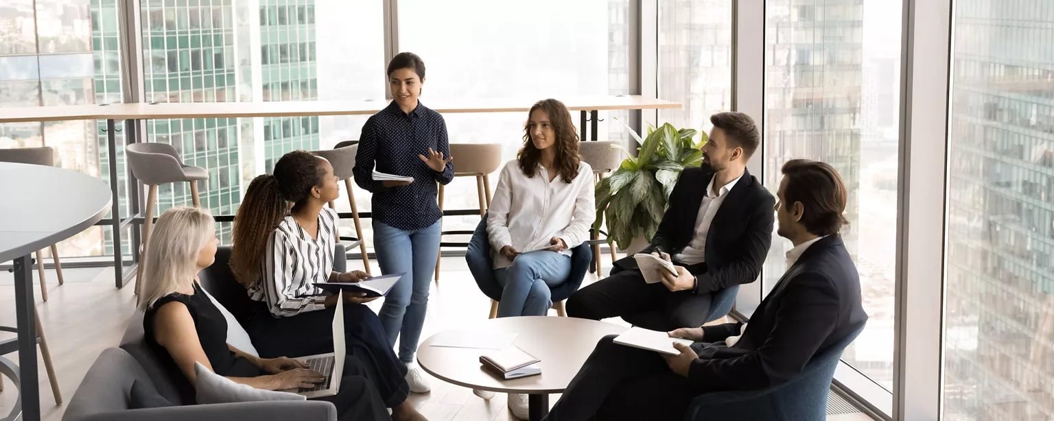 A female leader in a dark button-down shirt stands in the middle of a group of seated coworkers offering mentoring advice.