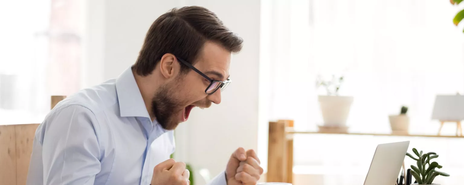 Young man with glasses sitting in front of a laptop looking happy and excited because he has received a great job offer.