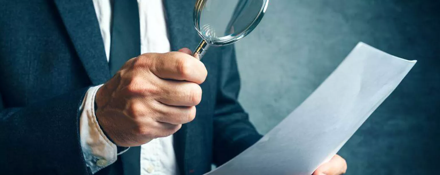 A man wearing a suit and tie uses a magnifying glass to scrutinize a piece of paper.