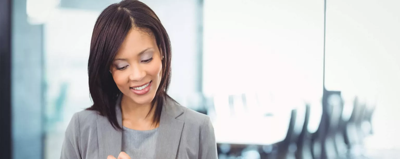 A businesswoman in a grey blazer and dress, standing in front of a conference room, tapping the screen on her smartphone and smiling.