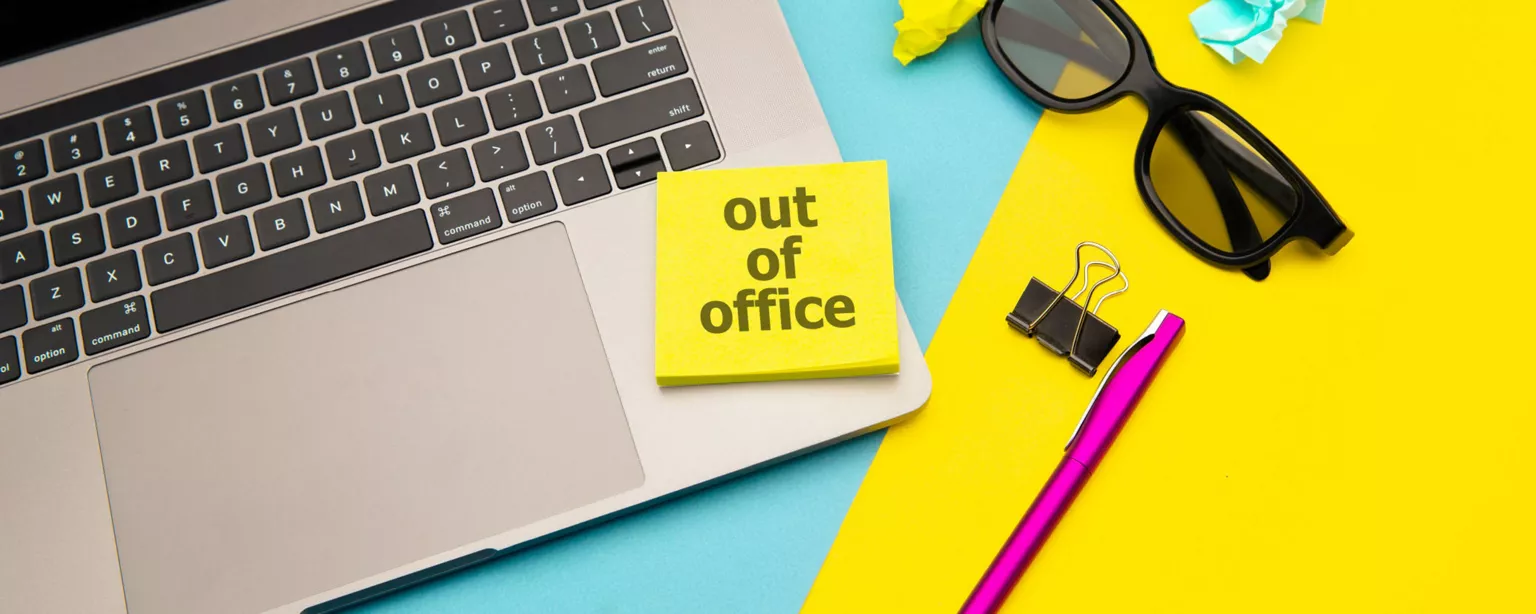 View of open laptop from above with post-it note on the keyboard reading "Out of Office" alongside sunglasses, pen and binder clip on top of a yellow folder