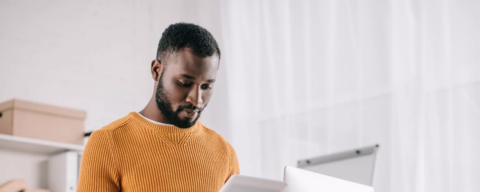 You man in a yellow sweater, using a tablet computer and standing in front of a desktop computer in a stylish office with white curtains.