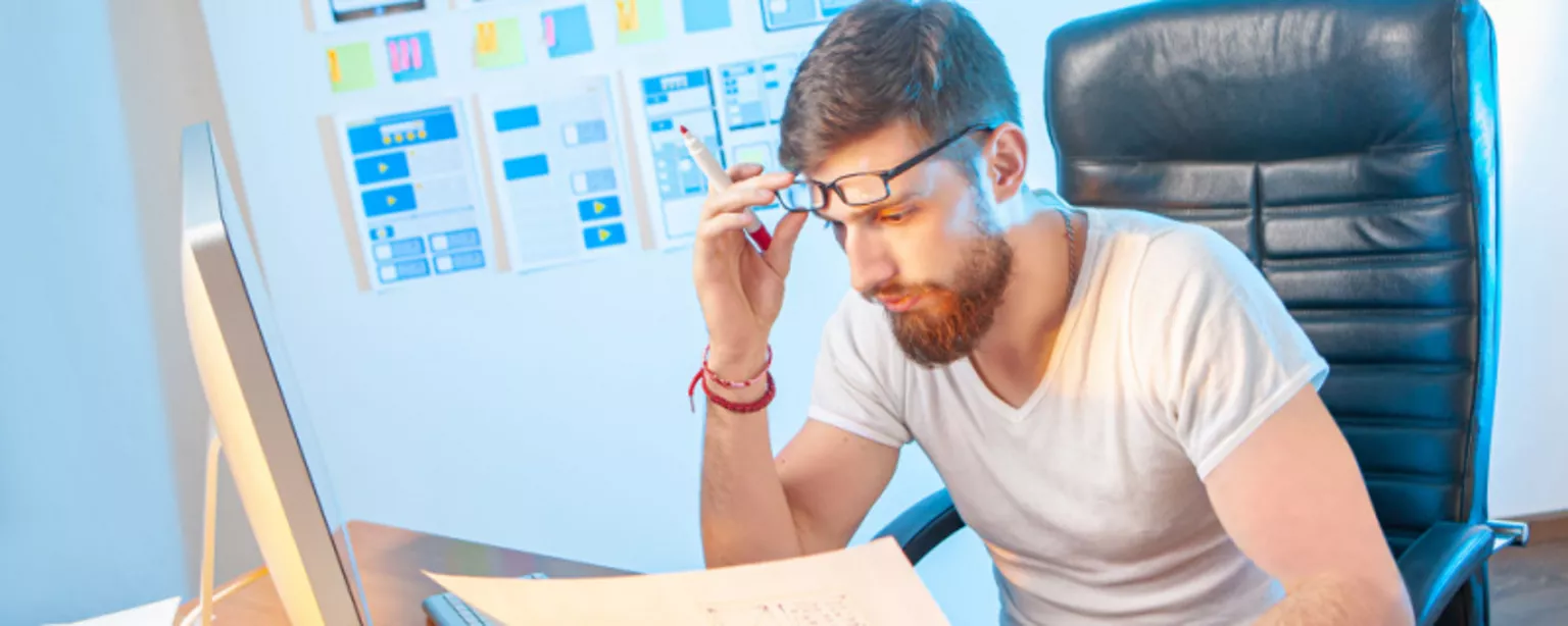 A young man with a beard and glasses, a mobile designer, sitting at a desk, closely examining a paper wireframe for a mobile application.