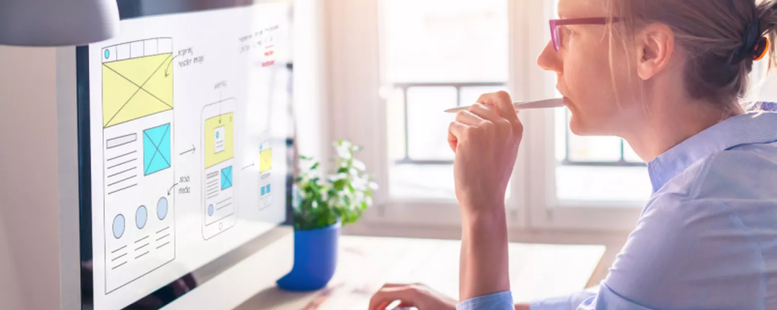 A woman in glasses, a web designer, contemplating a website layout design concept on her computer screen.