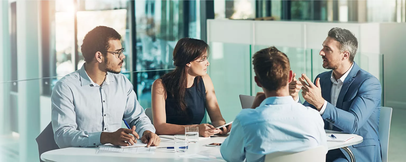 A group of male an female colleagues sit around a table listening to mentor speak.