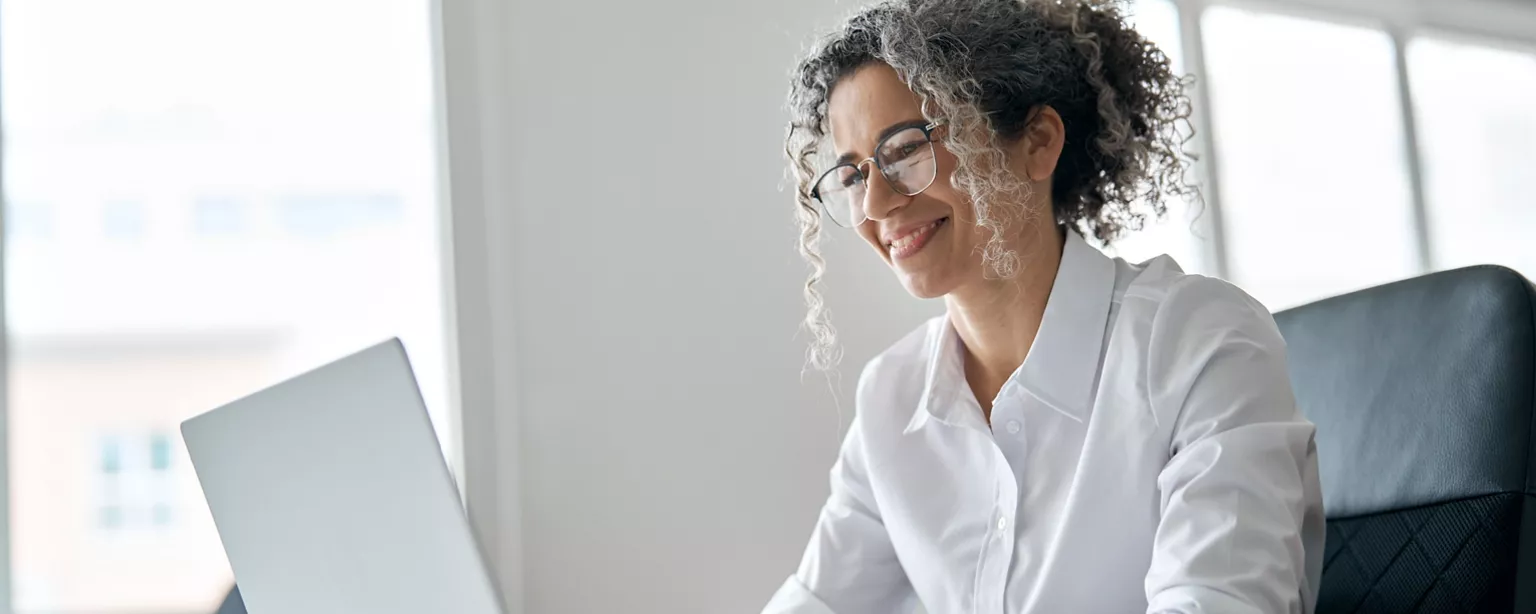 Female executive wearing glasses and a white shirt sits at a desk smiling at a computer.