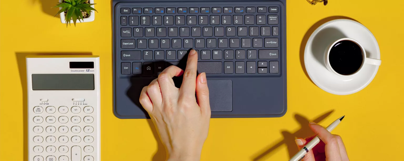 Laptop, calculator, coffee cup, eyeglasses and hands — one typing and one holding a pen — against bright a yellow background.