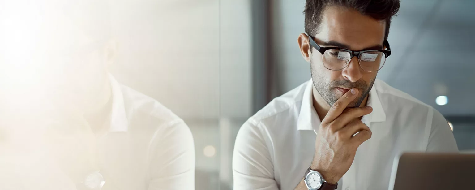 A man strikes a thoughtful pose as he looks at his laptop while writing an emailing requesting a raise.
