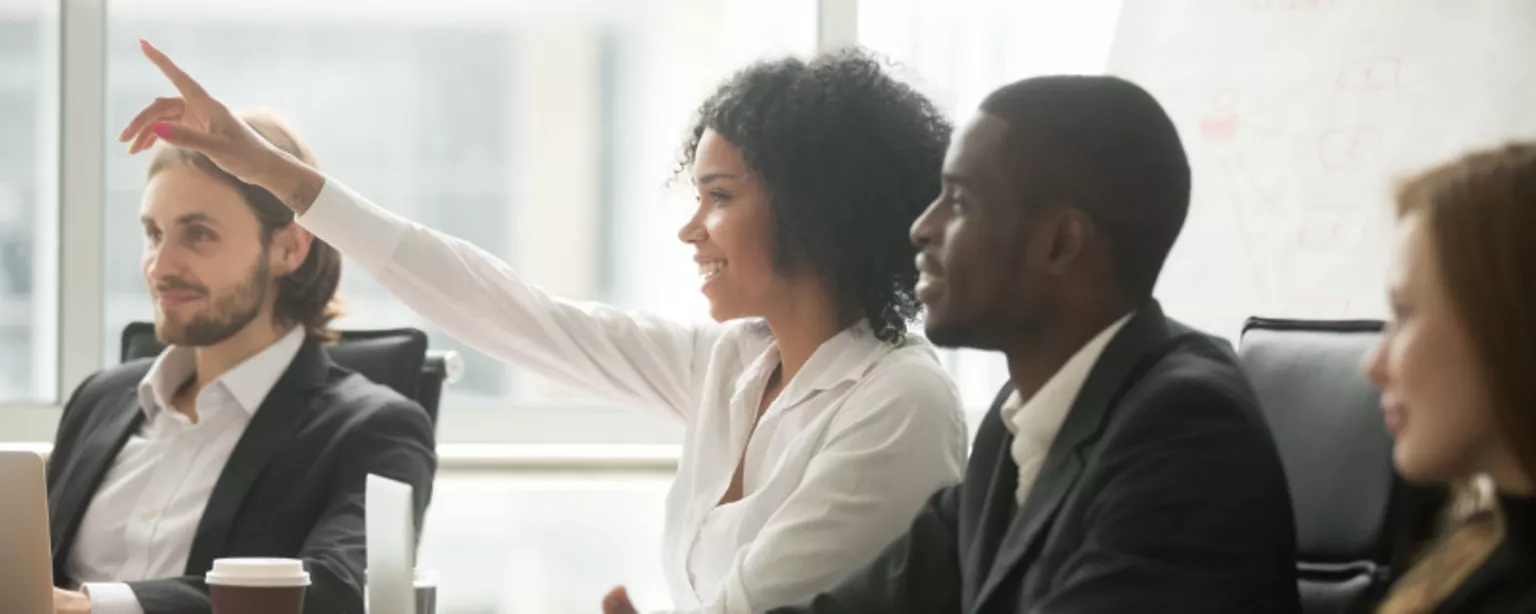 A young woman raises her hand to get a speaker's attention during an employee network group meeting that includes a diverse cross-section of coworkers.