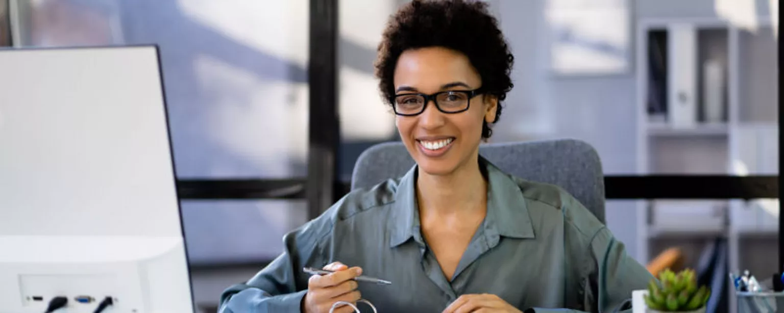 A payroll professional smiling and sitting at a desk with a calculator, binder and computer screen in front of them.