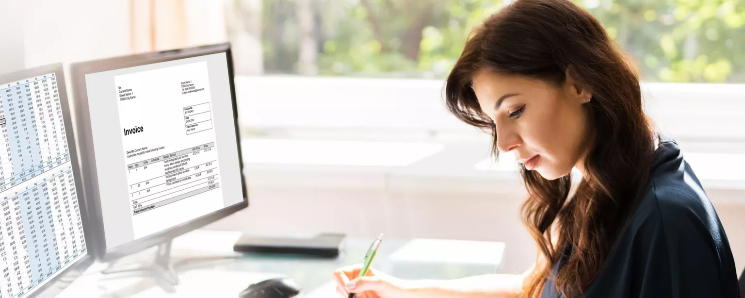 Woman sitting at a desk by a window using a calculator and writing in a binder. In front of her are two computer screens, one showing a spreadsheet and one showing a document that says "Invoice."