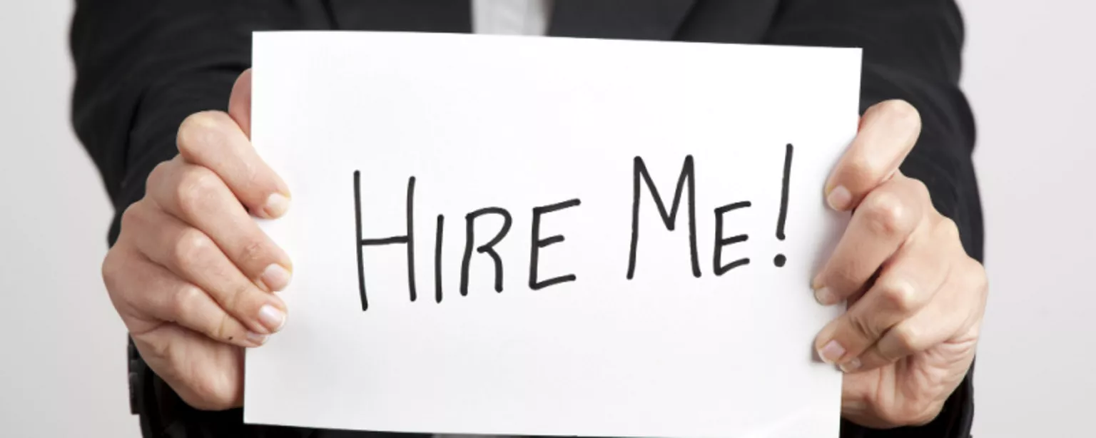 Hands hold a white sign with the words "Hire Me!" written in black marker.
