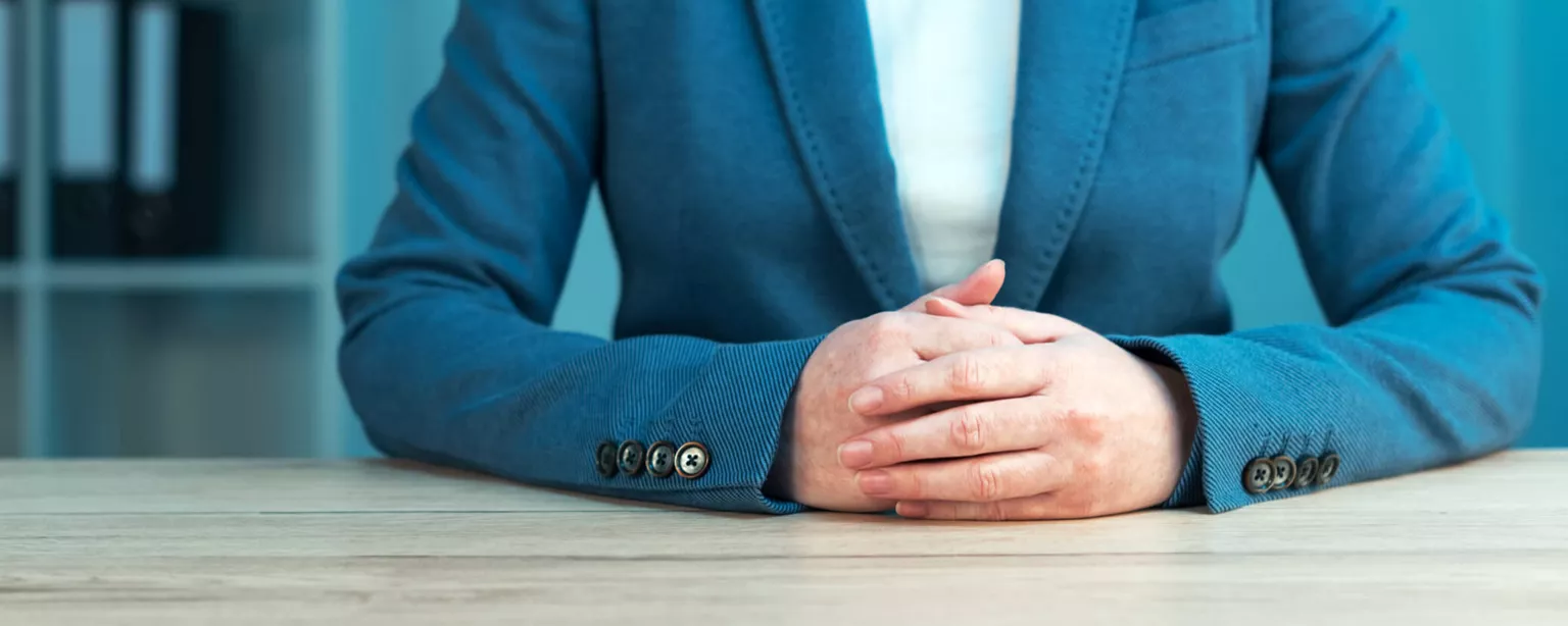 Woman in a blue business suit sitting at table, hands clasped. Her face is not visible.