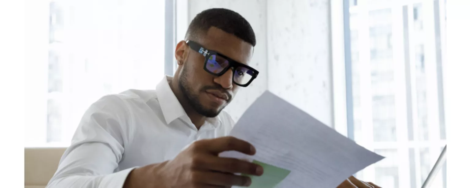 Young executive in stylish glasses and a white shirt, looking at a document while sitting in front of a laptop in a corporate office.