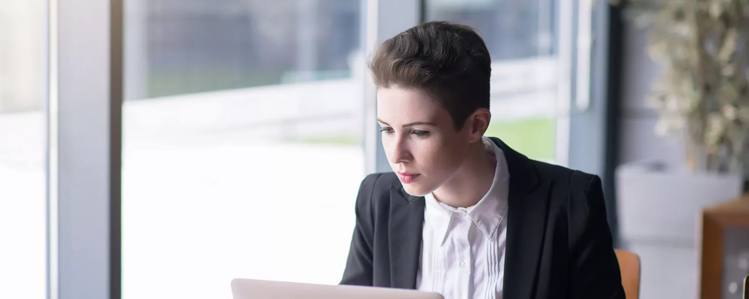 A young woman with short hair and business attire sits at a table and types at her laptop with her smartphone nearby.