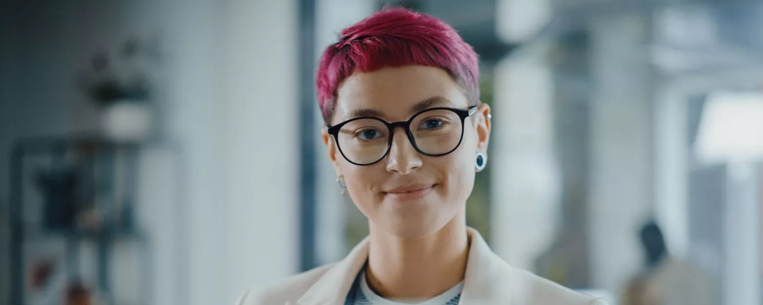 A smiling, young woman with short pink hair and glasses, wearing a white jacket and standing in front of a laptop.