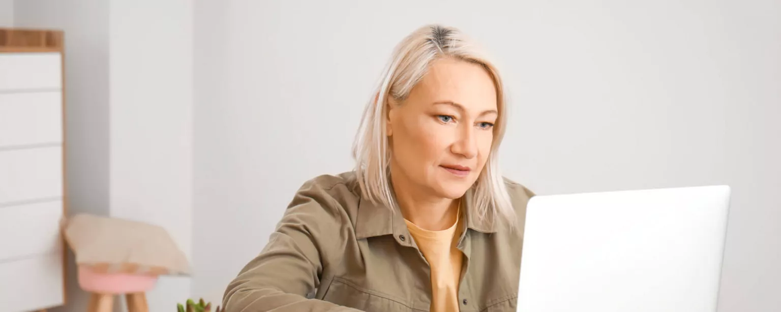 A woman with gray hair and a khaki shirt looking at a laptop in a home office.
