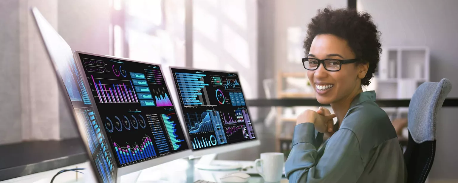 A woman who is a data scientist is smiling as she sits at her desk in front of a computer screens showing views of data and analytics.