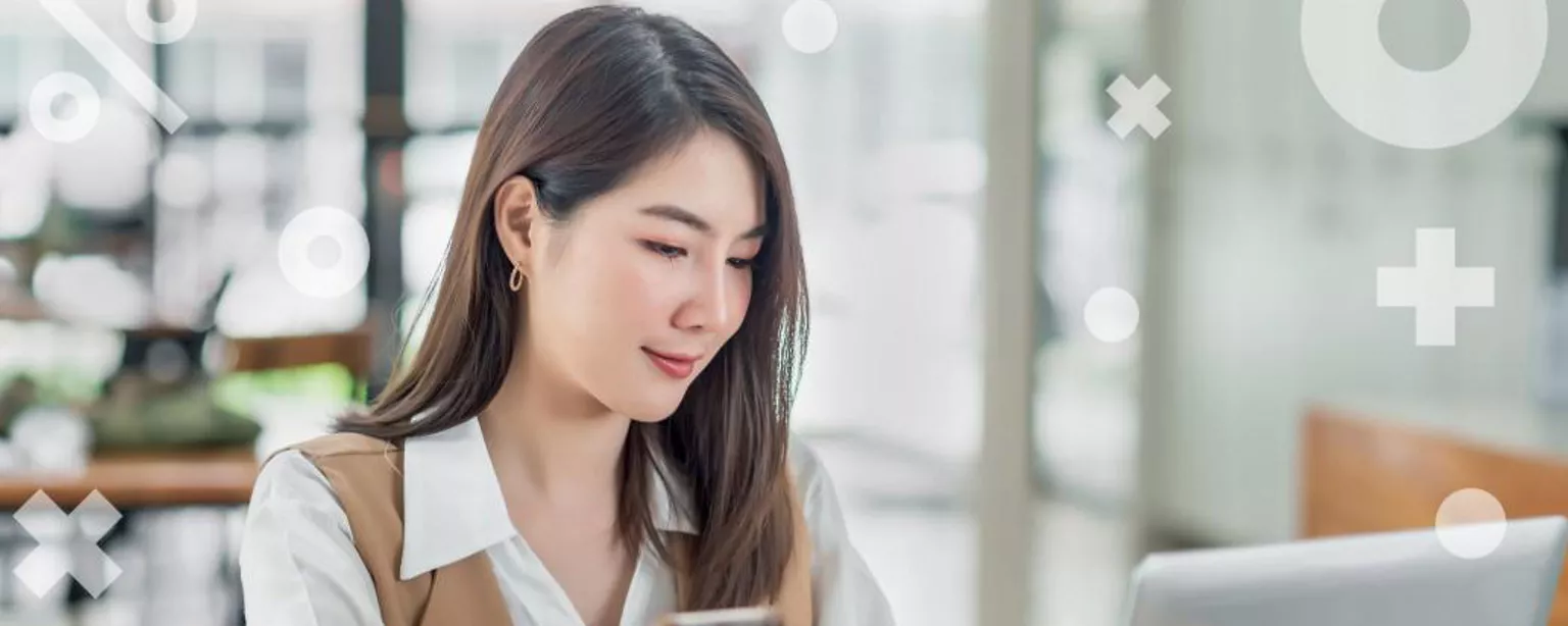 Jeune femme en gilet beige et chemise blanche, assise à un bureau, regardant l'écran d'un ordinateur portable et souriant.