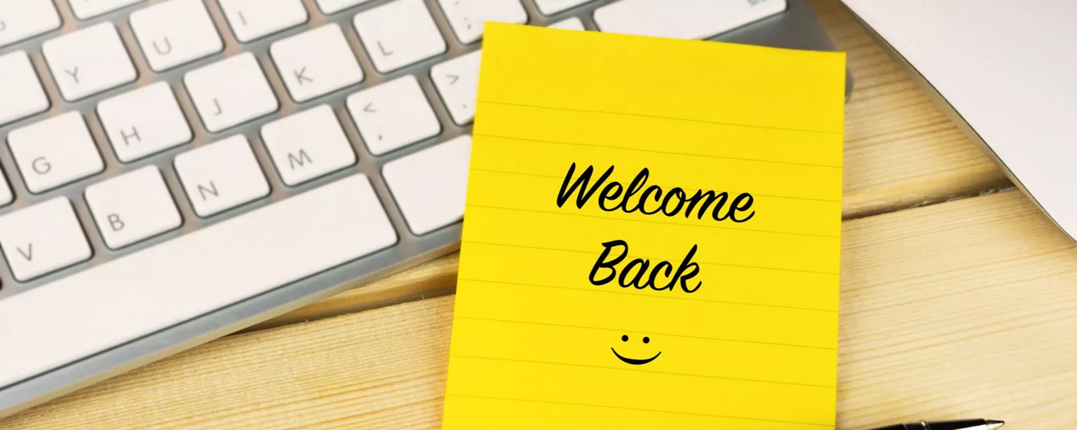 A yellow sticky note that says "Welcome Back" with a smiley face; the note is laying next to a computer keyboard, mouse and pen.
