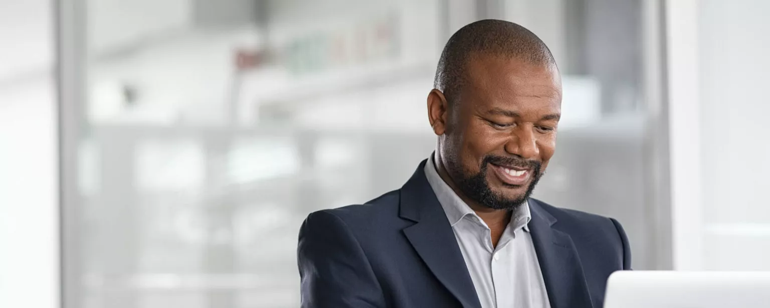 A businessman sitting at a desk, working on a laptop and smiling.