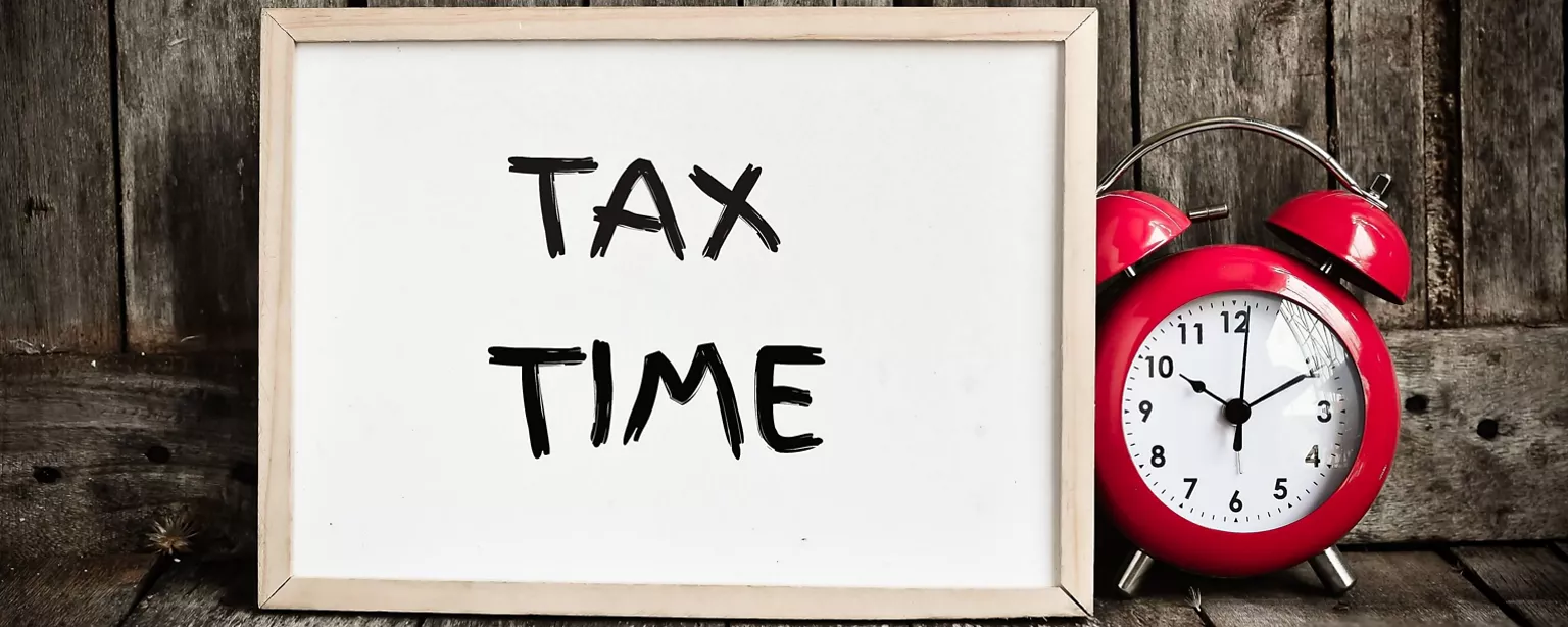 Red alarm clock on a wooden floor next to a white board that says, "TAX TIME" and is leaning against a wooden wall.