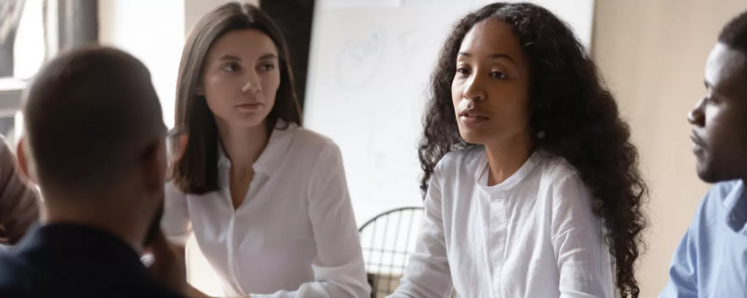 A female manager conveys compassion as she discusses layoffs with a group of employees in an office setting.